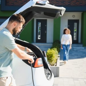 Homeowner connecting charging cable to electric car in front of modern house, embracing sustainable transportation