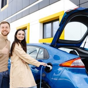 Young couple man and woman traveling by electric car having stop at charging station. Girlfriend holding power cable supply in hand while boyfriend gesturing finger up