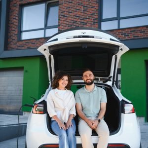 Happy couple sitting in open trunk of electric car, charging at home, enjoying sustainable lifestyle