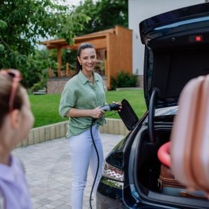 Happy mother holding power supply cable and charging their electric car, rest of family putting suitcases in a car trunk, preparating for holidays.